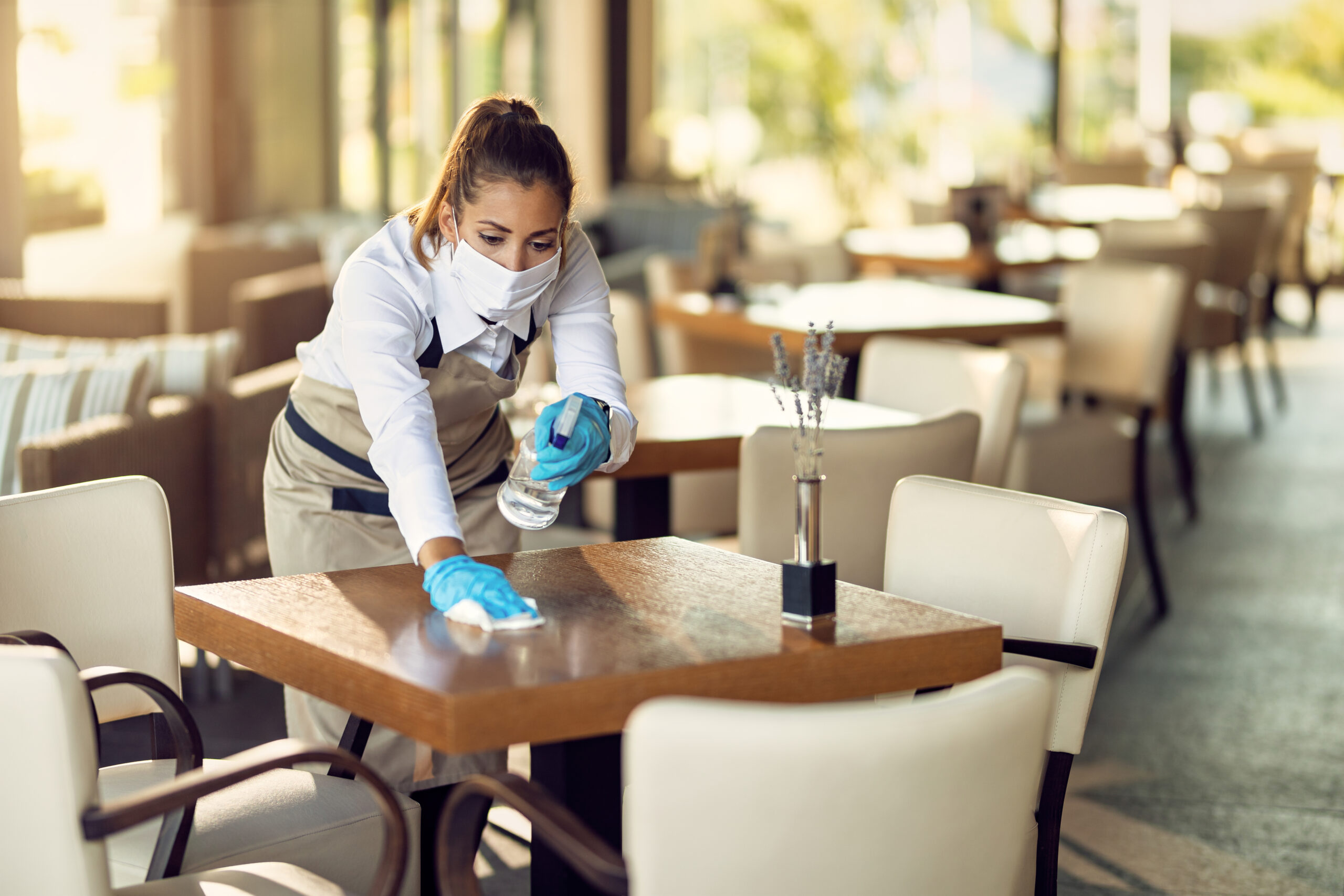 Waitress With A Face Mask And Gloves Cleaning Tables With Disinf